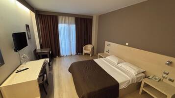 Una habitación de hotel con cama, mesita de noche, silla, escritorio, televisor y cortinas.