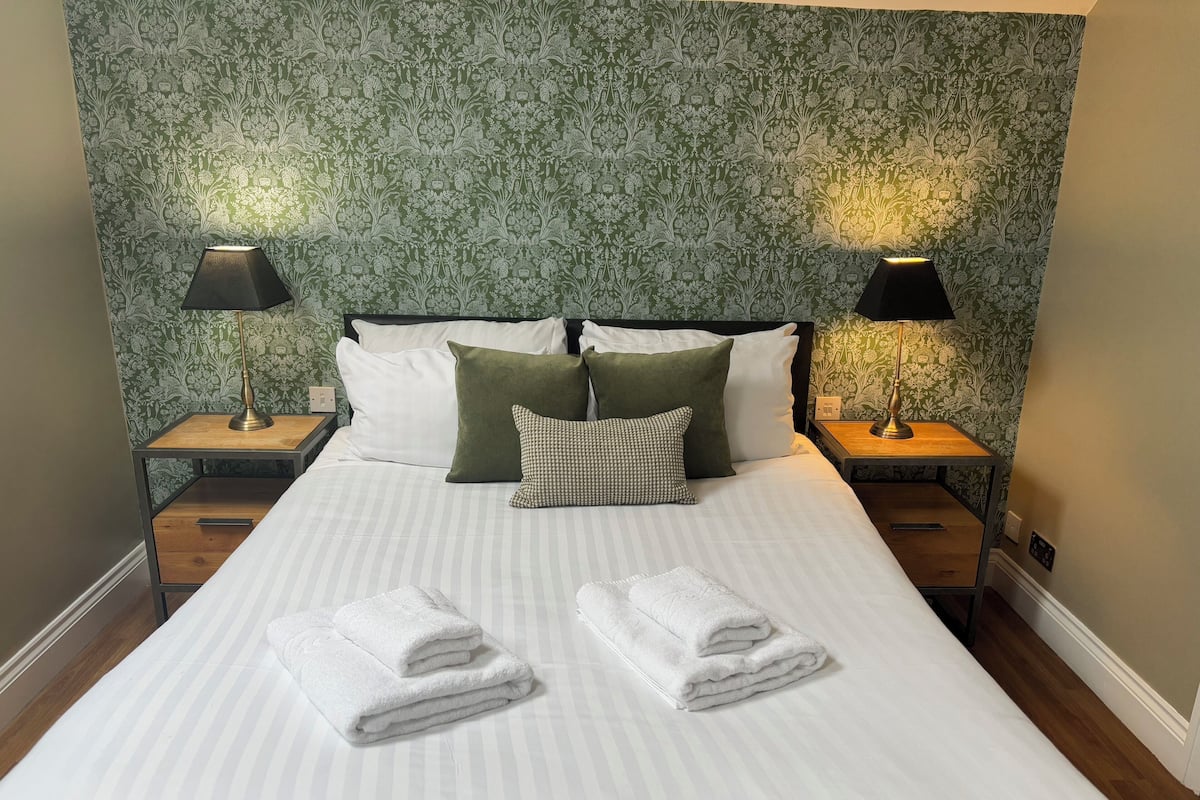 Cama bem arrumada com almofadas, duas luminárias de cabeceira e uma parede com padrão floral.