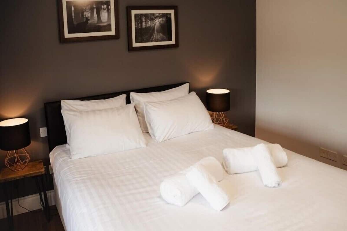 Cama bem arrumada com lençóis e almofadas brancas, duas luminárias de cabeceira e duas fotos emolduradas na parede.