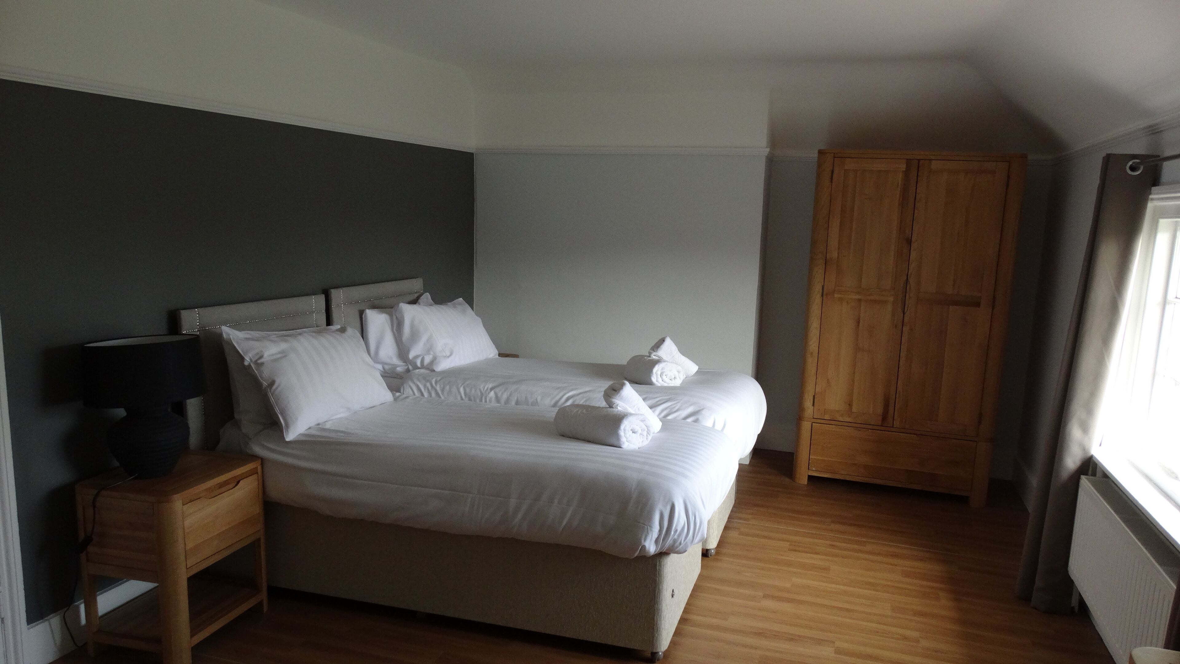 Una cama bien hecha con sábanas y toallas blancas, una mesita de noche de madera con una lámpara, y un armario de madera en una habitación con suelo de madera y una pared de acento gris.