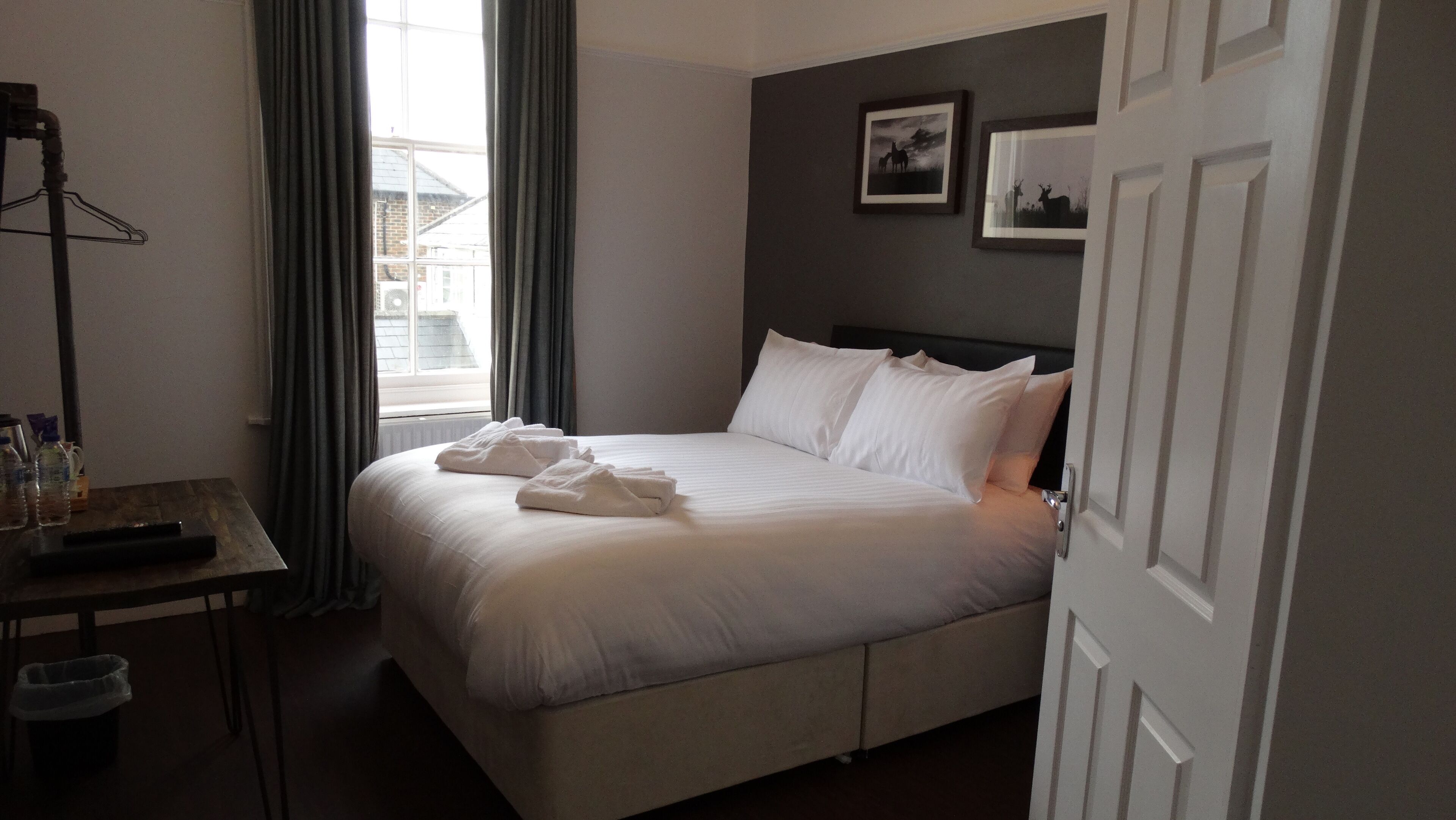 A neatly made bed with white linens and towels, a bedside table with a remote control, and a window with curtains.