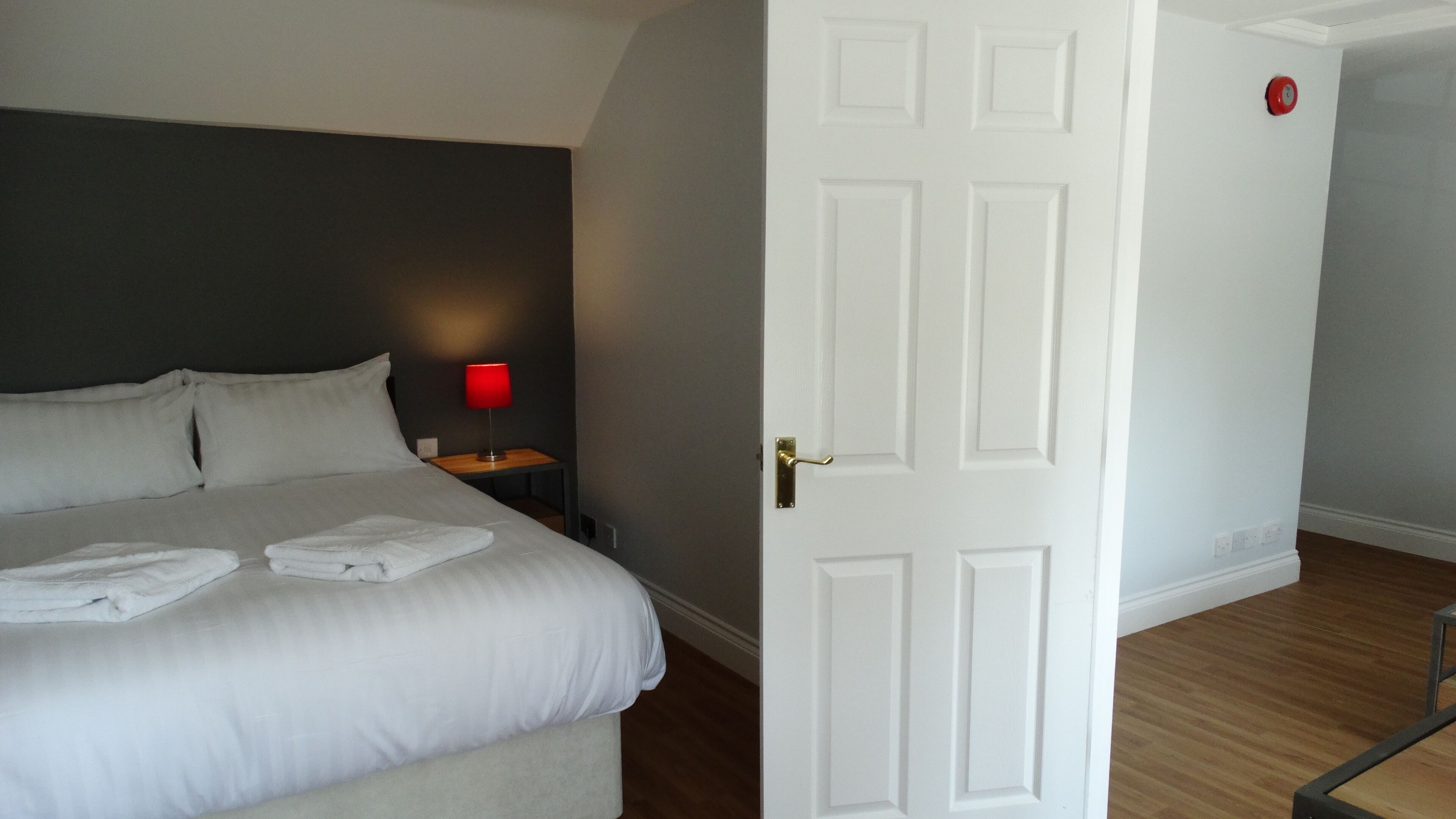 A hotel room with a bed, a nightstand with a red lamp, and a white door.