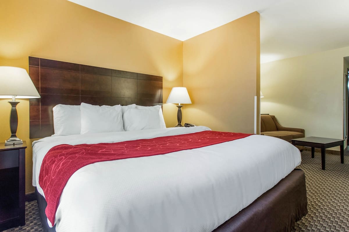 A hotel room with a large bed, two bedside lamps, a red throw blanket, a brown headboard, and a small table with a chair.