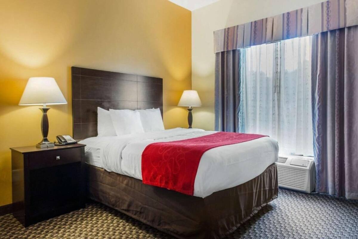 A hotel room with a bed, two bedside tables with lamps, a window with curtains, and a wall heater.