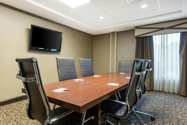 A conference room with a long table, six chairs, a flat-screen TV on the wall, and a large window with curtains.