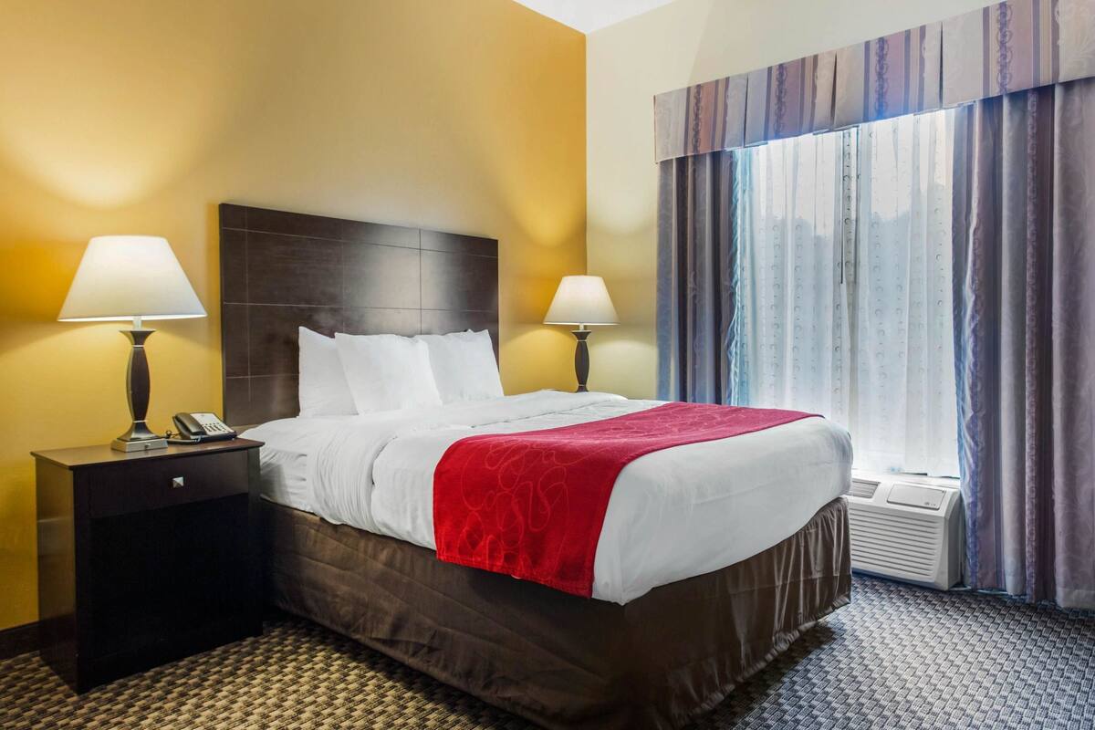 A hotel room with a large bed, two bedside lamps, a telephone, a window with curtains, and a wall-mounted air conditioning unit.