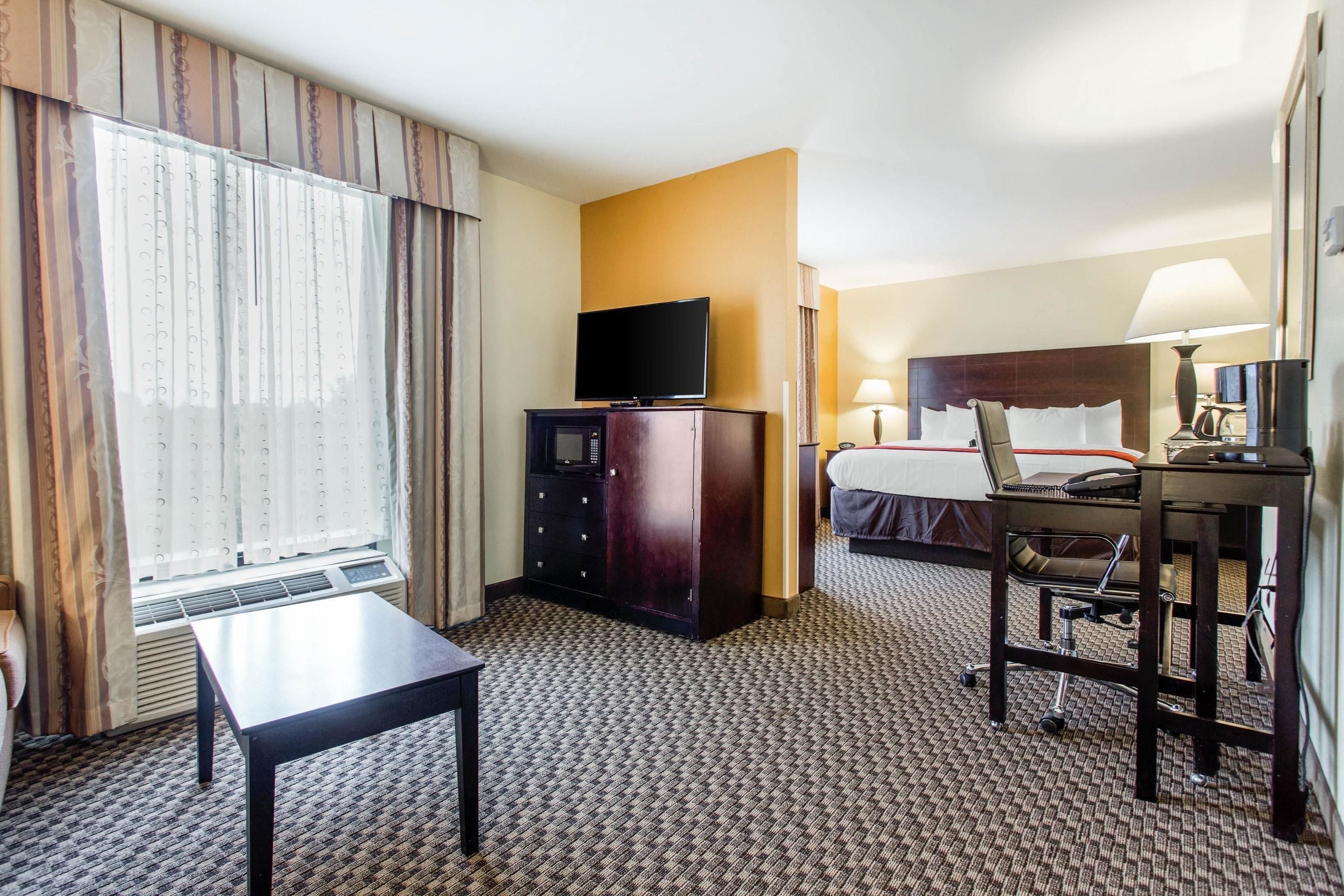 A hotel room with a bed, a desk with a chair, a television, a desk lamp, and a window with curtains.