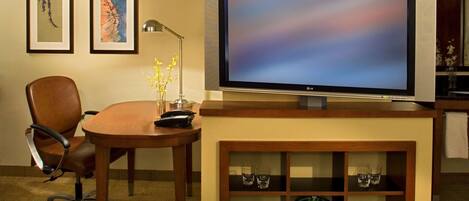 A hotel room with a wooden desk, a chair, a television on a stand, and two framed artworks on the wall.