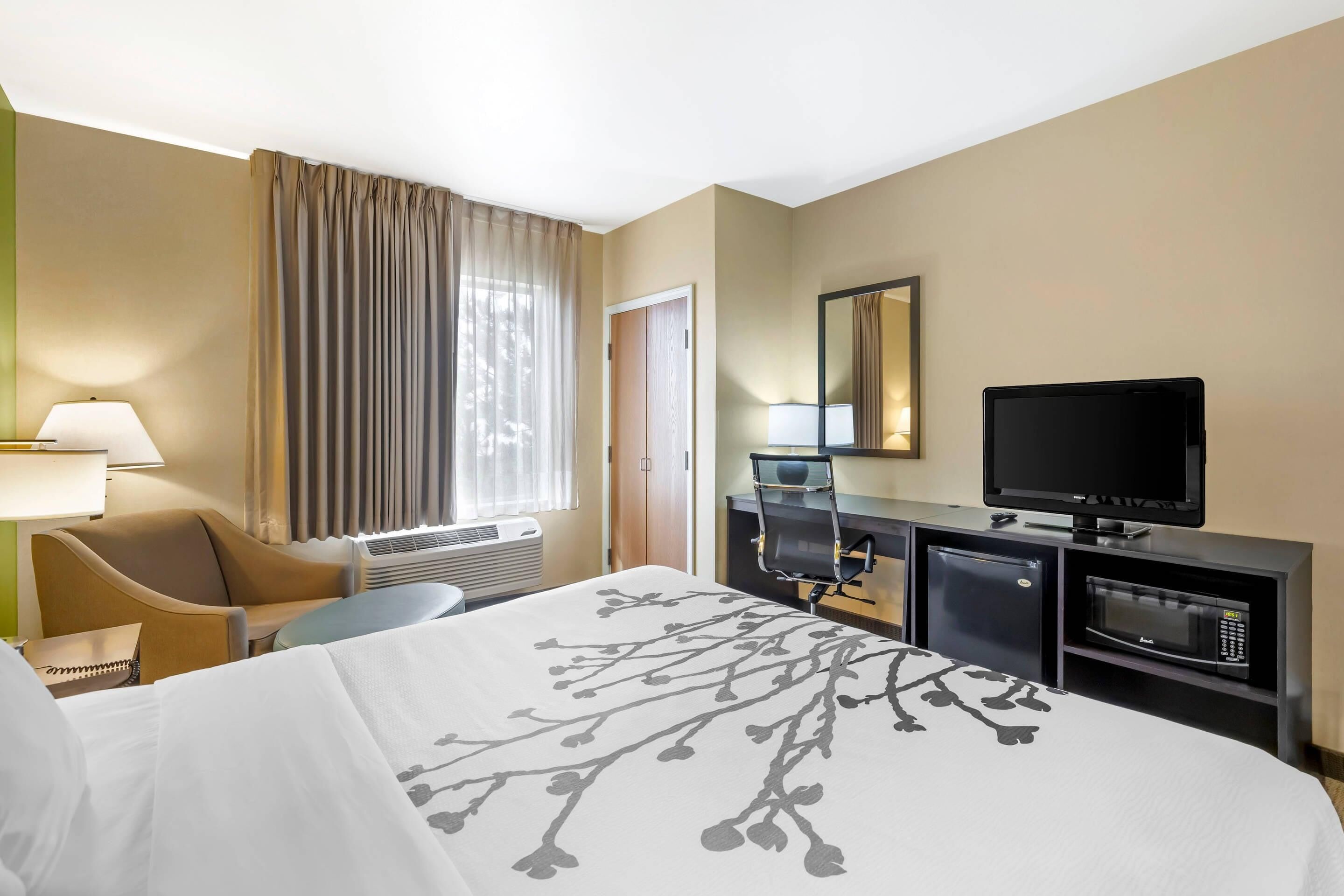 Ein Hotelzimmer mit einem Bett, einem Schreibtisch mit Fernseher, einem Sessel, einer Schreibtischlampe und einem Fenster mit Vorhängen.