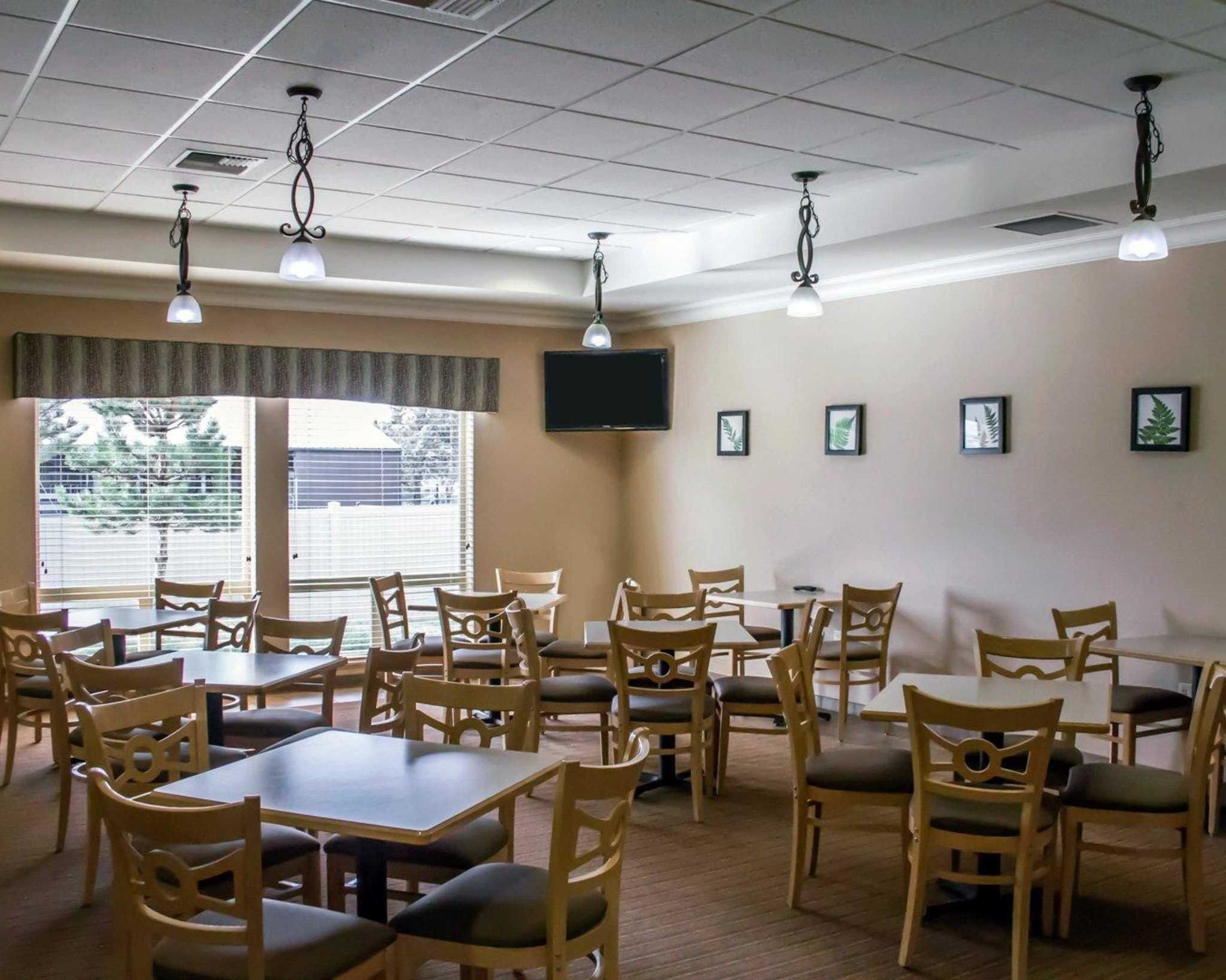 A dining area with multiple tables and chairs, a flat-screen TV mounted on the wall, and large windows with blinds.
