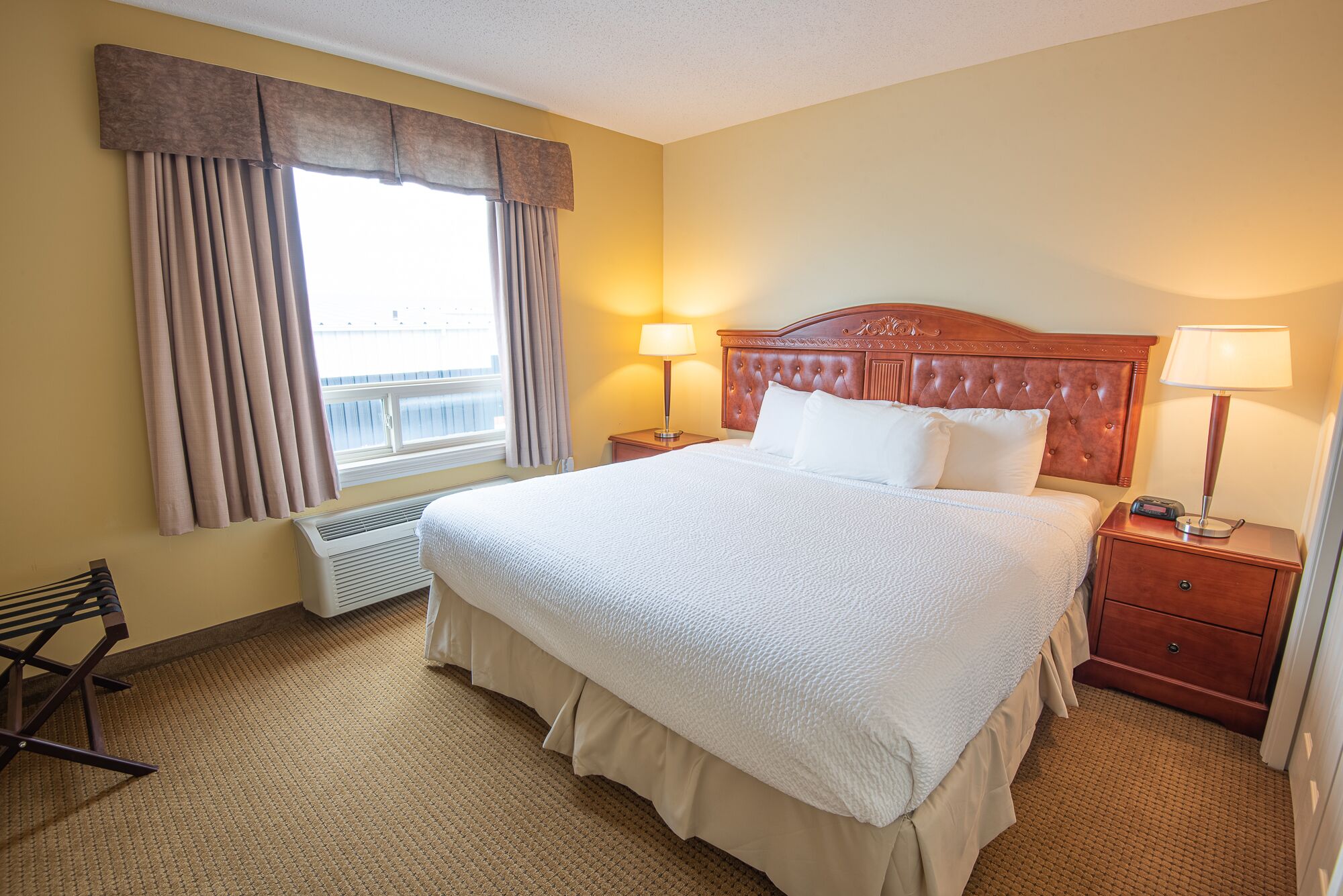 A hotel room with a large bed, two bedside tables, a window with curtains, a radiator, and a bench.