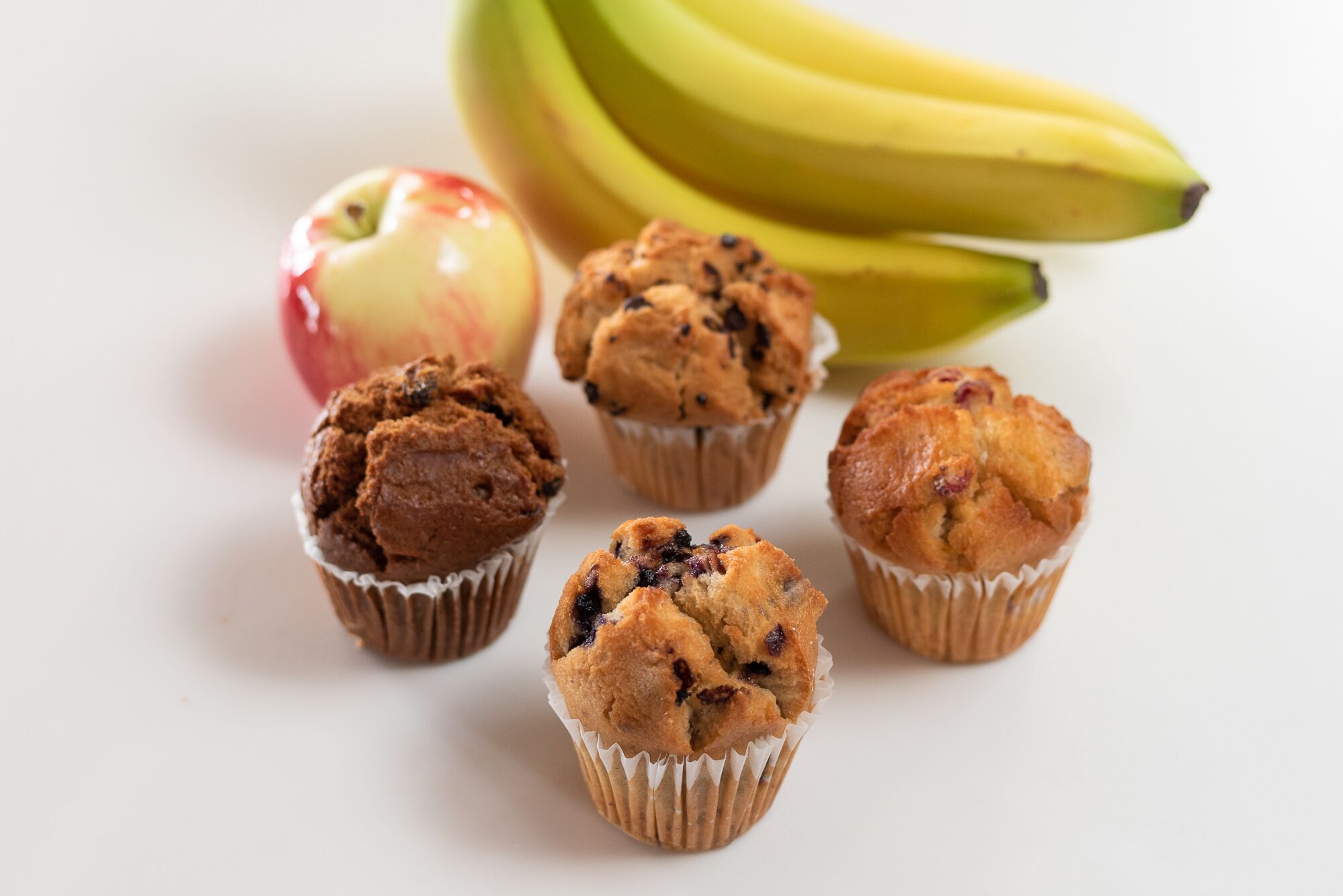 Een wit oppervlak met vijf muffins in papieren bekers, een rode appel en een tros bananen.