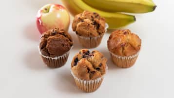 Een wit oppervlak met vijf muffins in papieren bekers, een rode appel en een tros bananen.