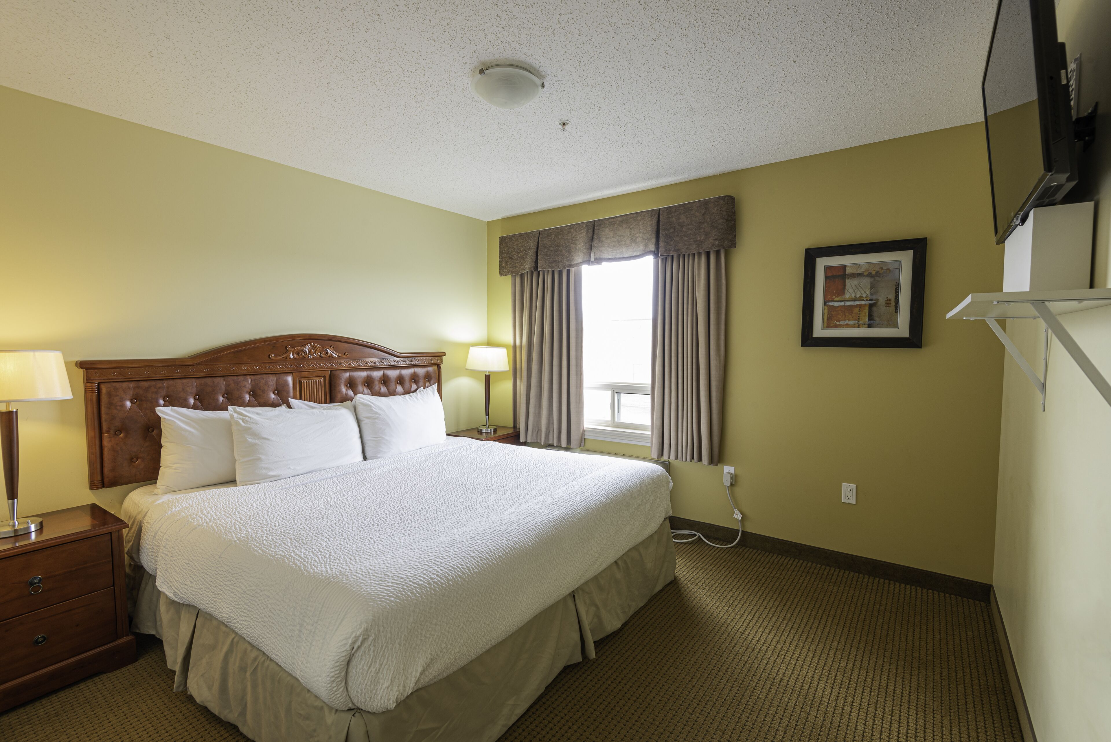A hotel room with a large bed, two bedside tables, a nightstand, a lamp, a window with curtains, a framed picture on the wall, and a television mounted on the wall.