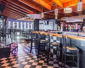 Uma área de bar com piso xadrez, banquetas e um balcão com diversos frascos e copos.