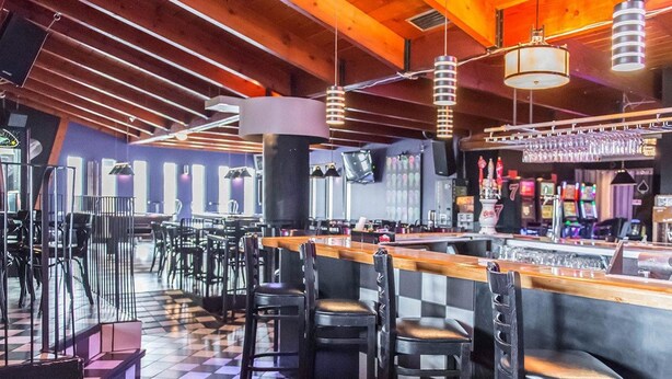 Zona de bar con suelo a cuadros, taburetes negros y barra con una variedad de botellas y vasos.
