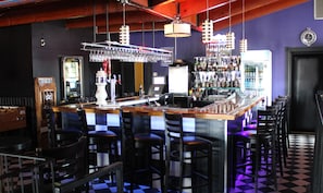Una zona de bar con suelo a cuadros, taburetes y una barra con enfriador de vino y varias botellas.