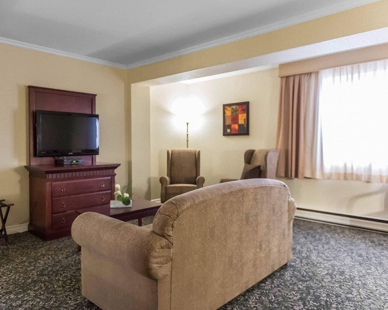 Quarto de hotel com sofá, televisão sobre um armário de madeira, uma cadeira e uma janela grande com cortinas.