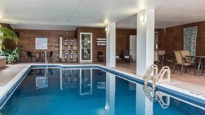 Una piscina interior con agua cristalina, rodeada de paredes de madera y un suelo de baldosas. Hay sillas y mesas alrededor de la piscina, y se observa un cartel en la pared.