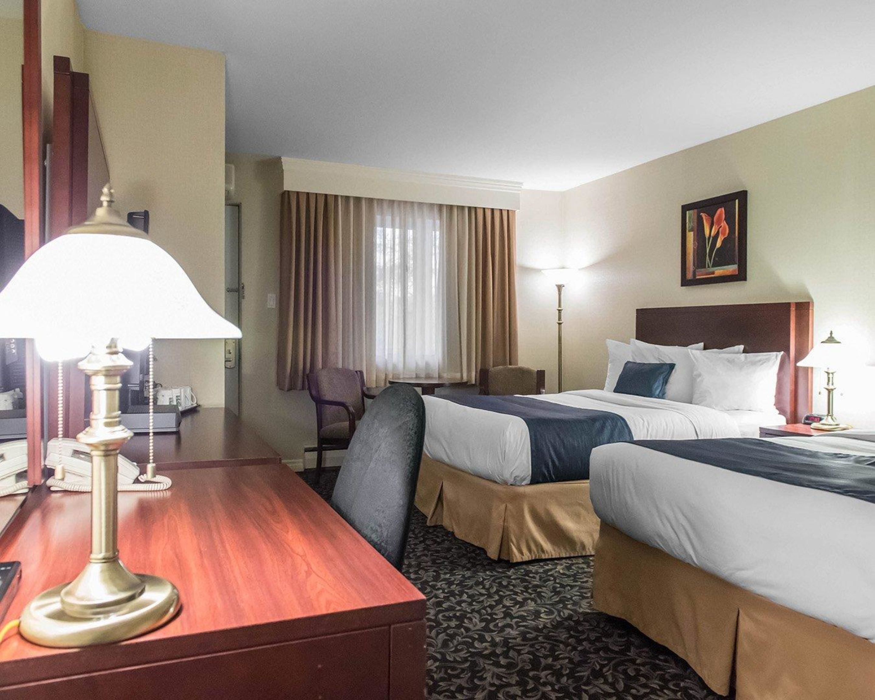 A hotel room with two beds, a desk with a lamp, a chair, a desk lamp, a painting on the wall, and a window with curtains.