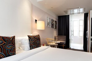 A hotel room with a bed, a chair, a small table, a lamp, and a sliding glass door leading to a balcony. - Le Placide Saint Germain des Pres (Paris)