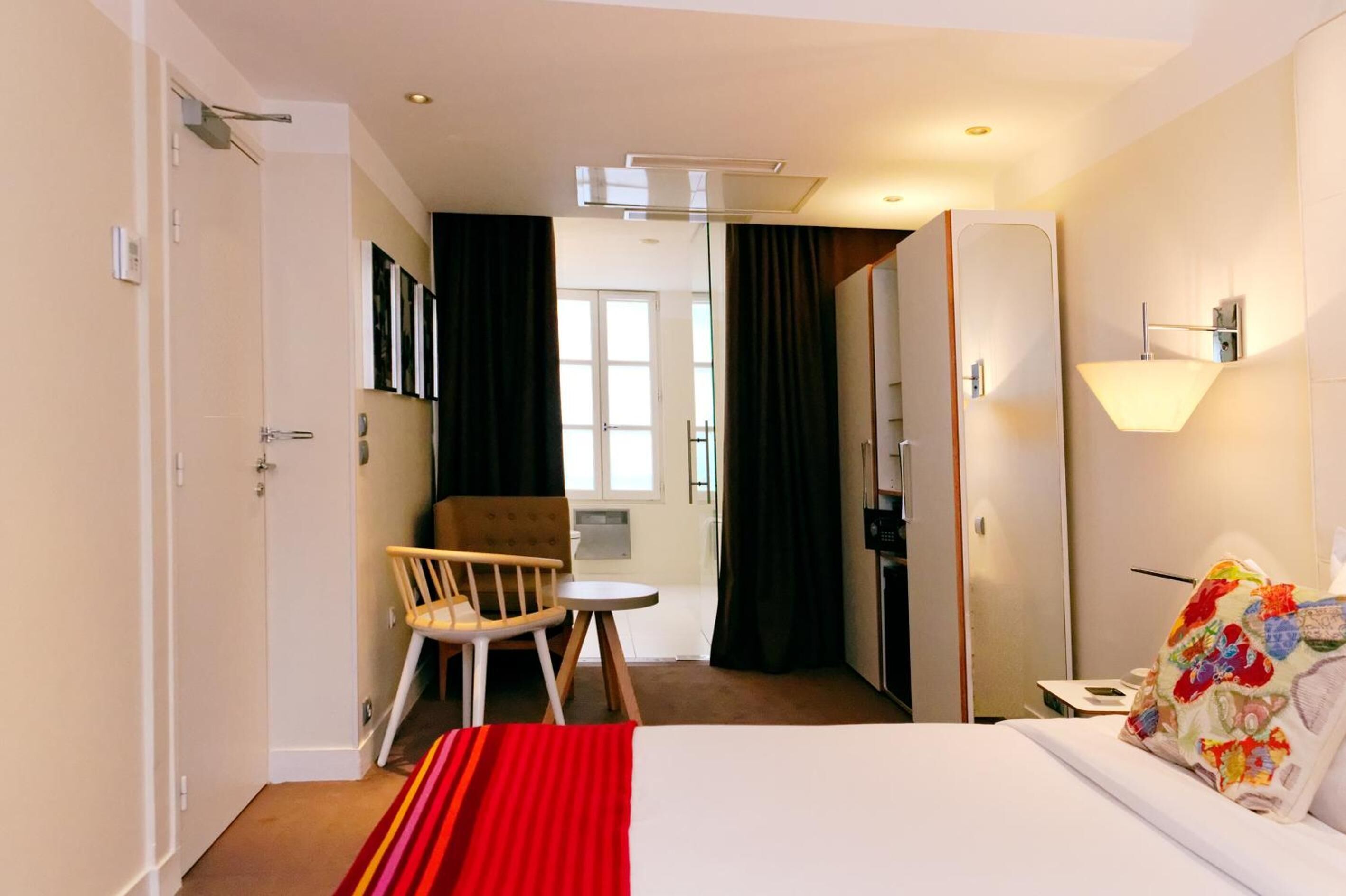 A hotel room with a bed, a chair, a small table, a lamp, and a view of the hallway.