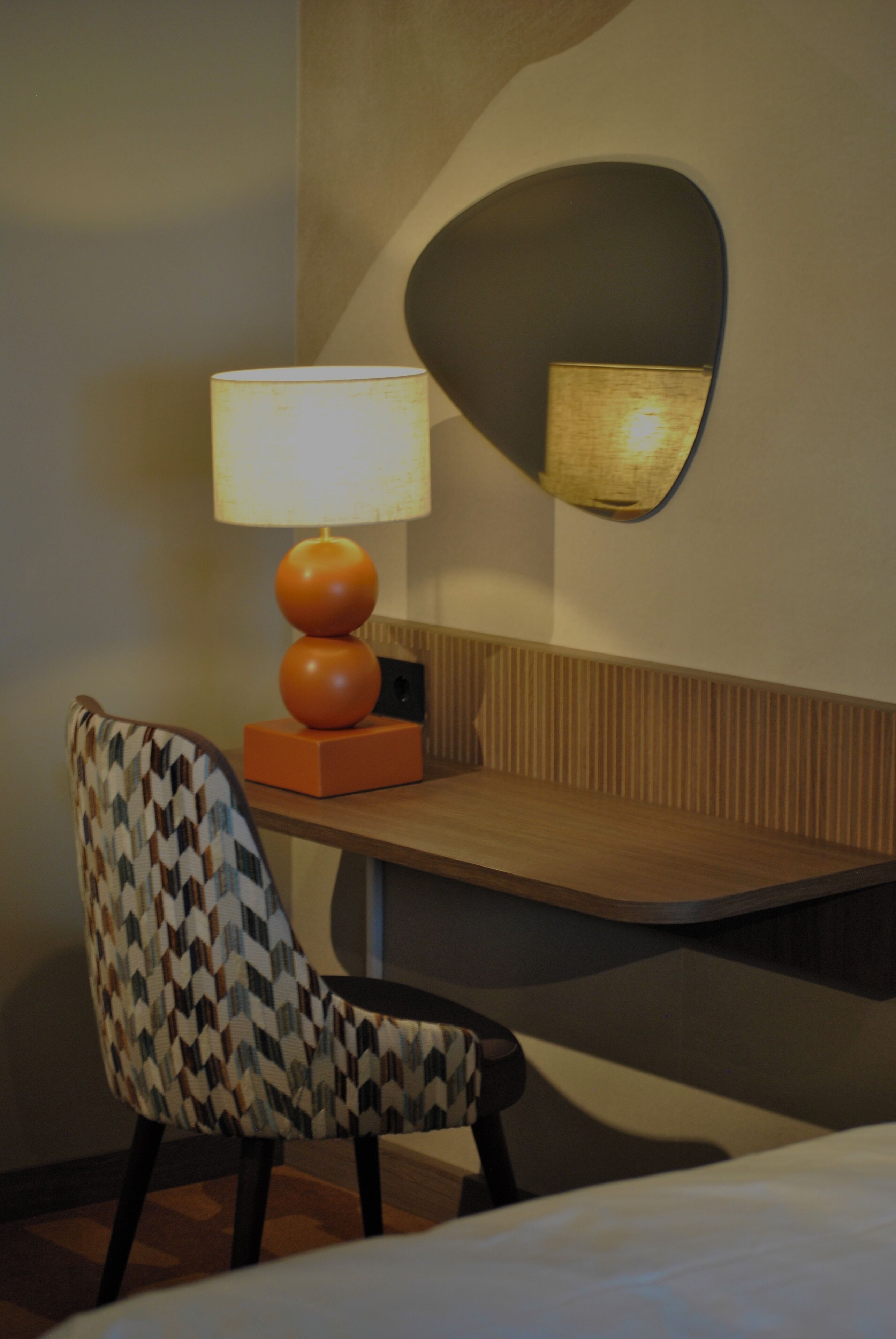 A hotel room with a bed, a chair, a table, a lamp, and a wall-mounted mirror.