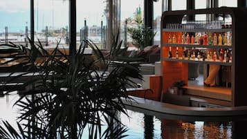 Zona de bar con una planta grande en primer plano, una barra con diferentes botellas y vistas al puerto deportivo a través de amplios ventanales.