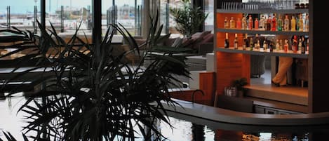 Un espace bar agrémenté d’une grande plante en pot au premier plan, d’un comptoir de bar où sont exposées diverses bouteilles, et d’une vue sur la marina, visible à travers de grandes fenêtres.