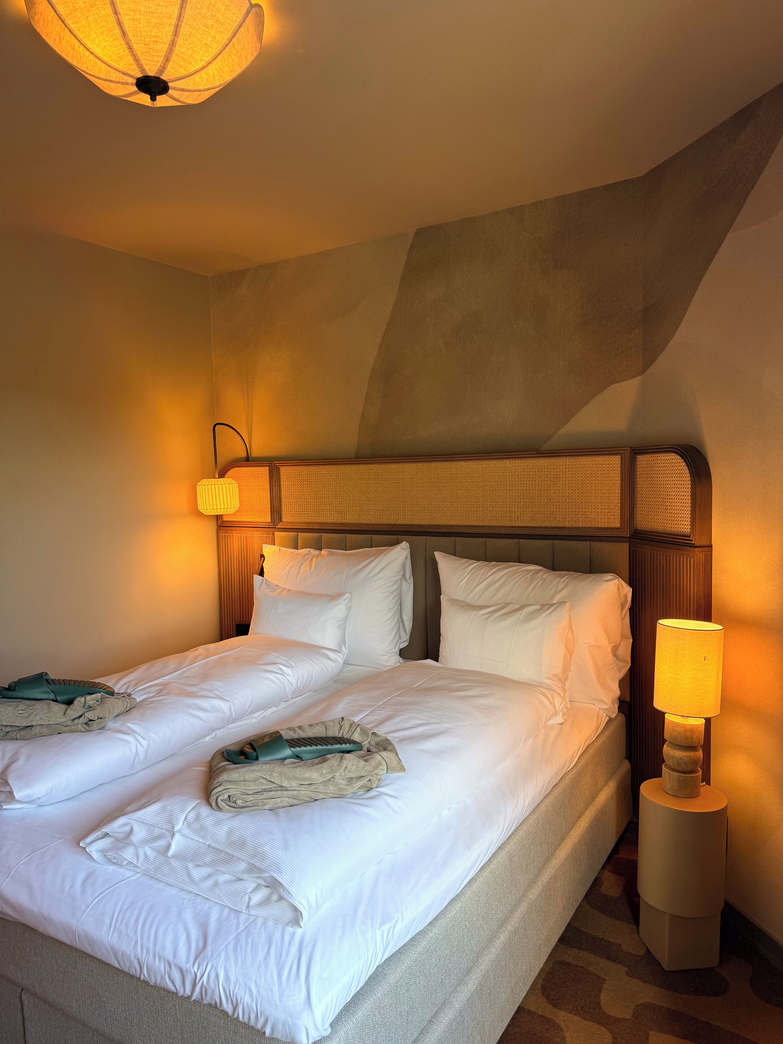 Cama doble con ropa de cama blanca, cabecera con panel texturizado, lámpara de noche y aplique de pared.