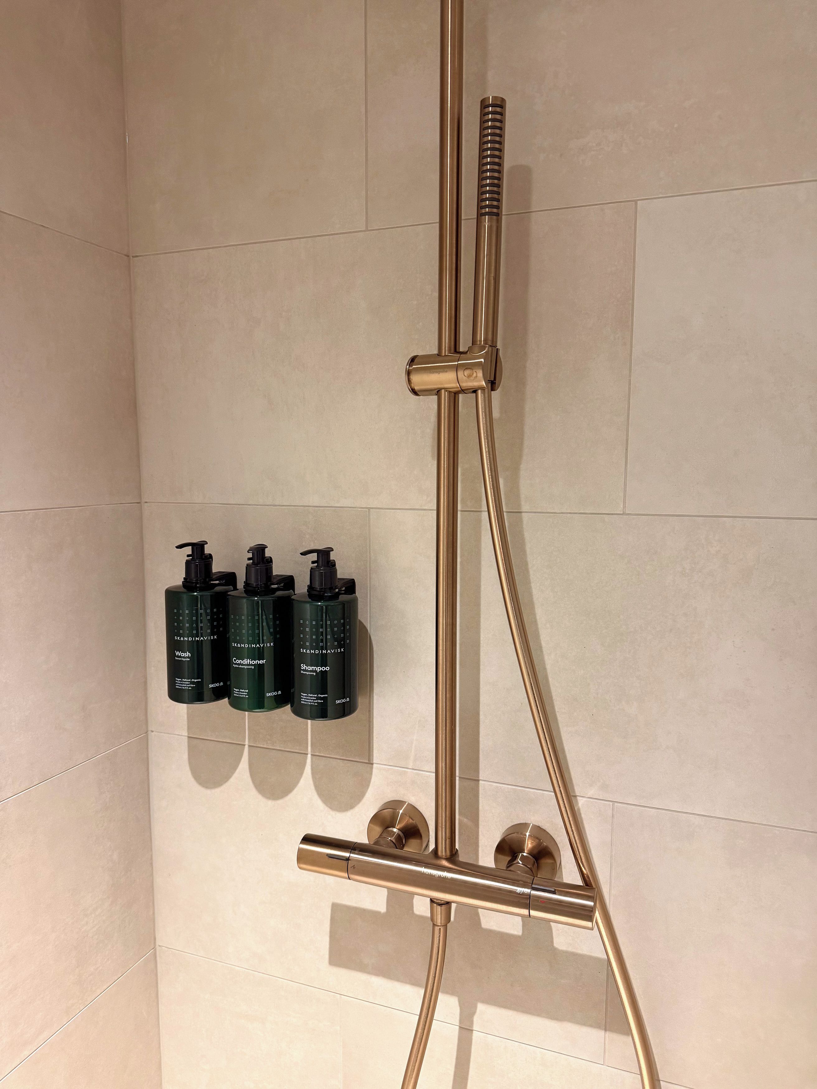Un baño con sistema de ducha color dorado y tres botellas de productos para el cuidado de la piel escandinavos.
