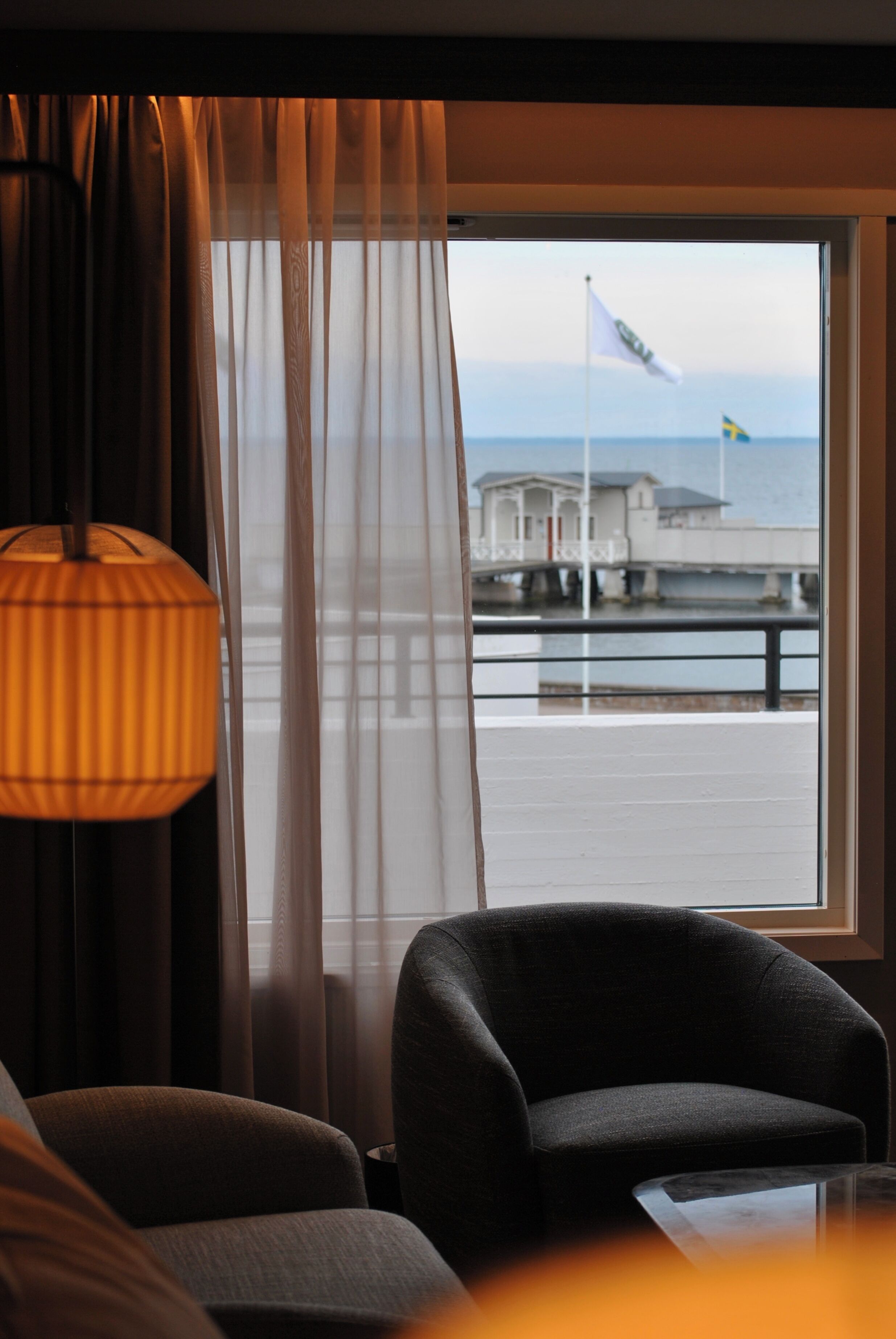 A room with a view of a pier and a flag, a lamp, and a chair.