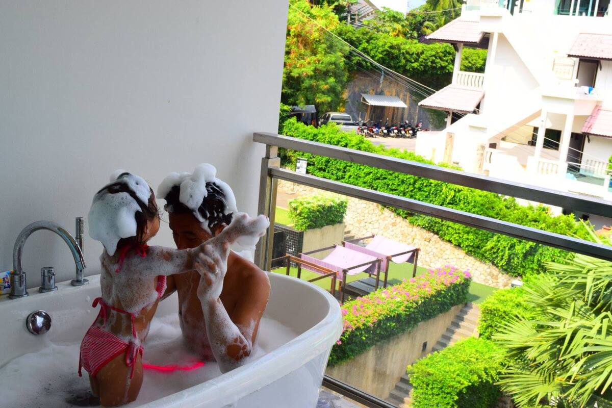 Una bañera con dos personas bañándose, un balcón con vistas a un jardín y edificios, y una pequeña mesa con sillas afuera.