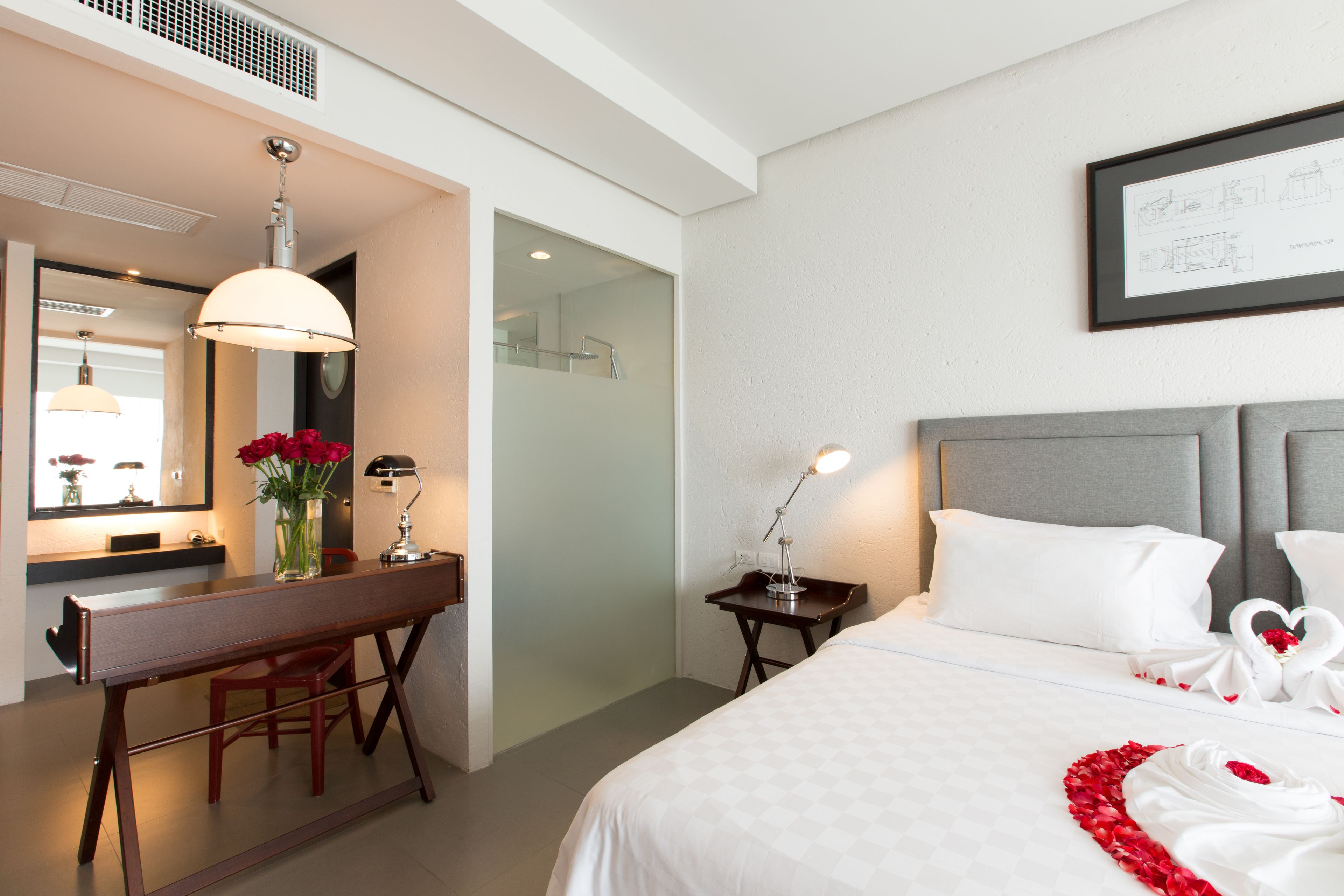 A hotel room with a bed, a desk, a chair, a mirror, and a bedside table with a lamp.