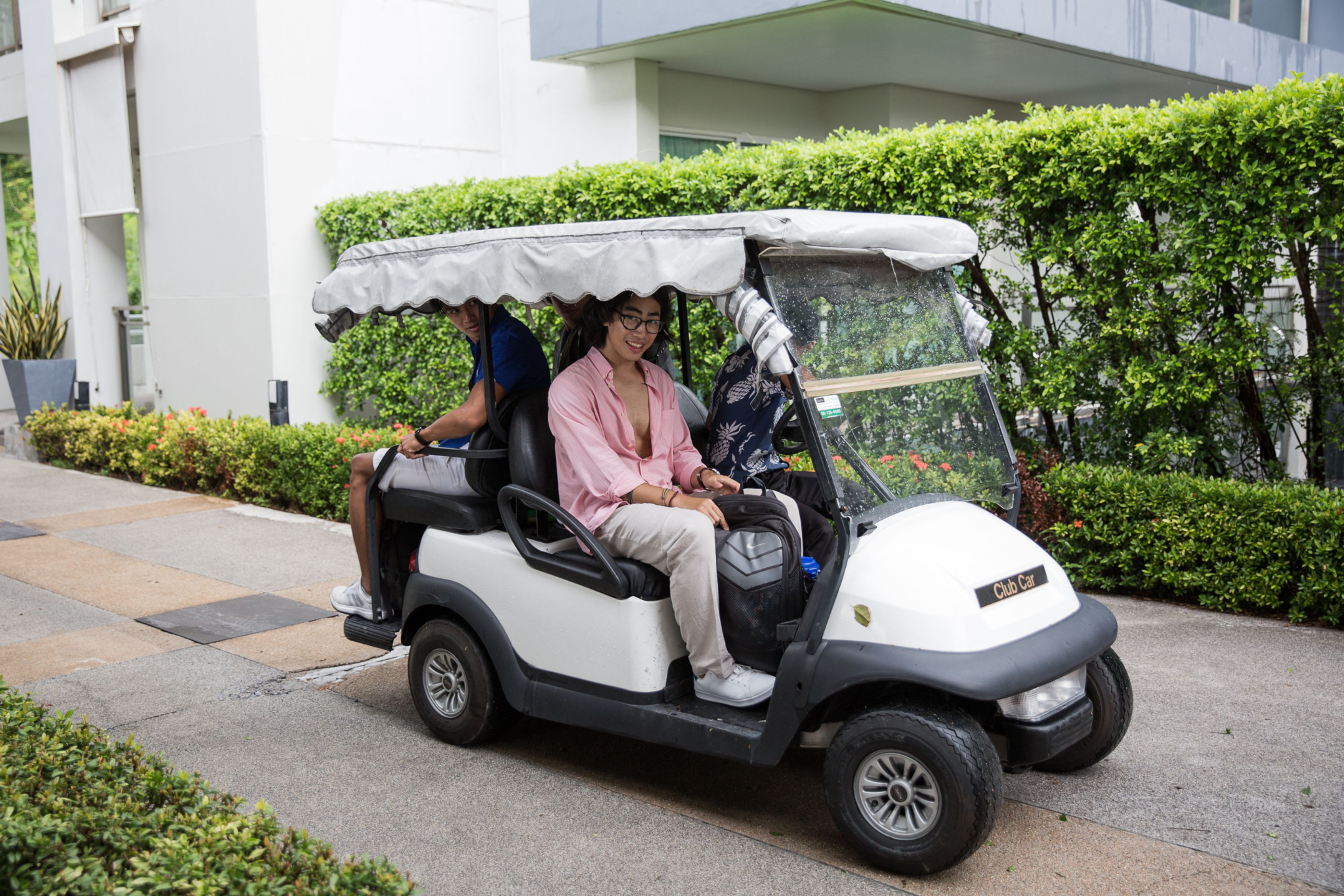 A golf cart with a canopy, parked on a paved path beside a building with a hedge.