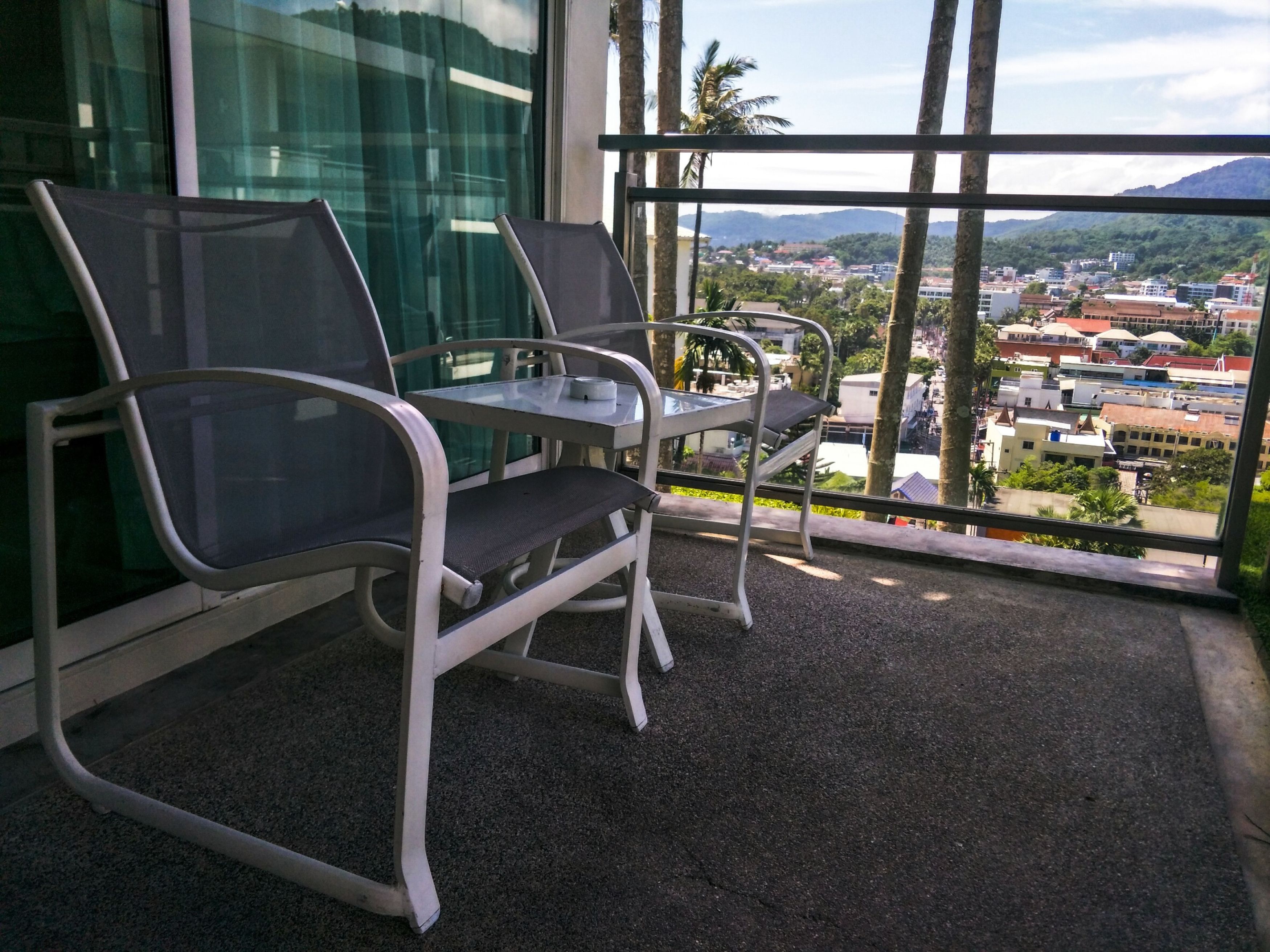 A balcony with two chairs and a table, overlooking a cityscape with buildings and mountains.