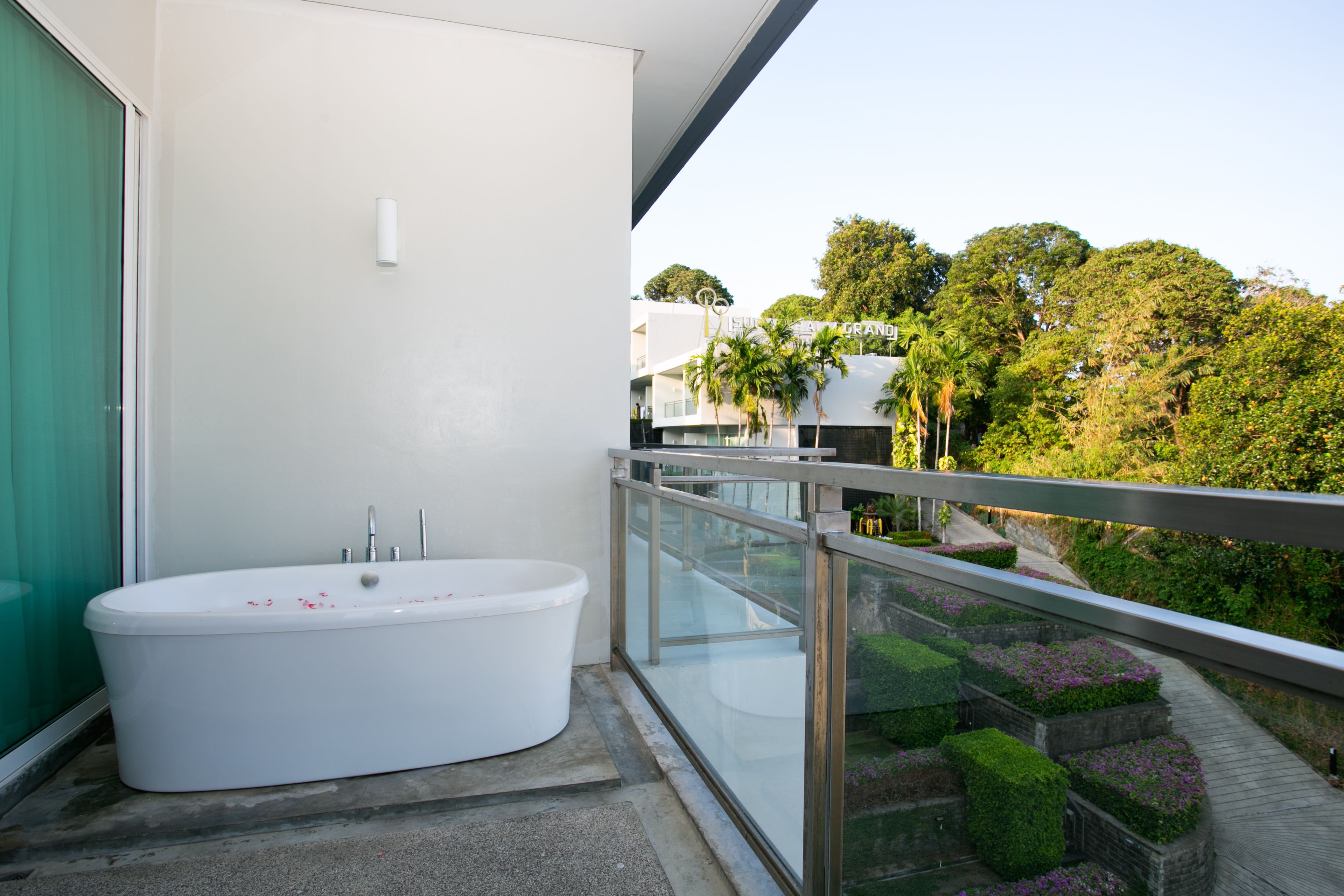 Une salle de bain moderne avec une baignoire sur pieds, une douche en verre et un balcon doté d’une rambarde en métal, offrant une vue sur un jardin aménagé et des arbres.