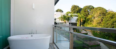 Une salle de bain moderne avec une baignoire sur pieds, une douche en verre et un balcon doté d’une rambarde en métal, offrant une vue sur un jardin aménagé et des arbres.