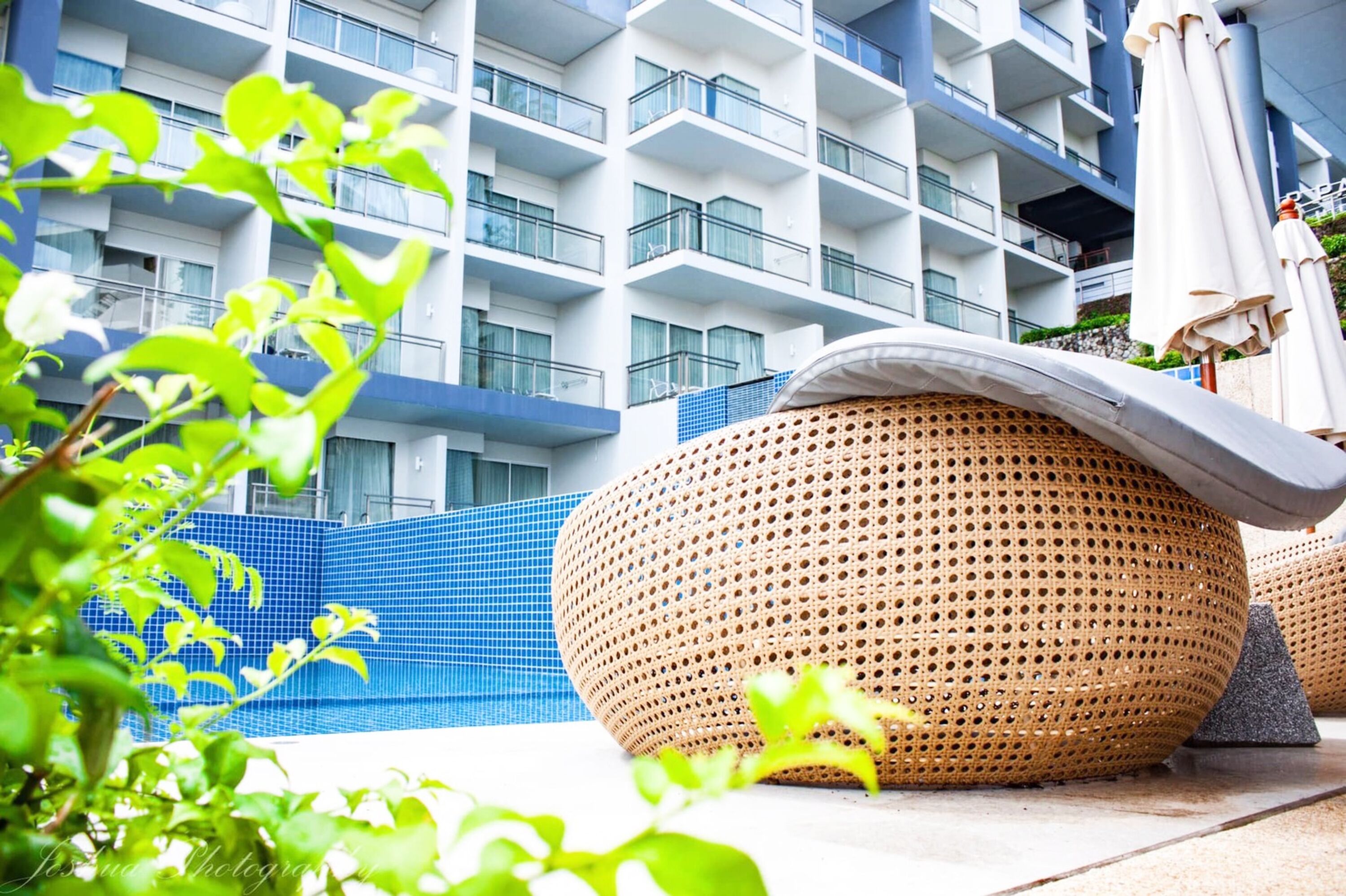 A modern hotel with a blue tiled pool area, large woven seating, and a multi-story building with balconies.