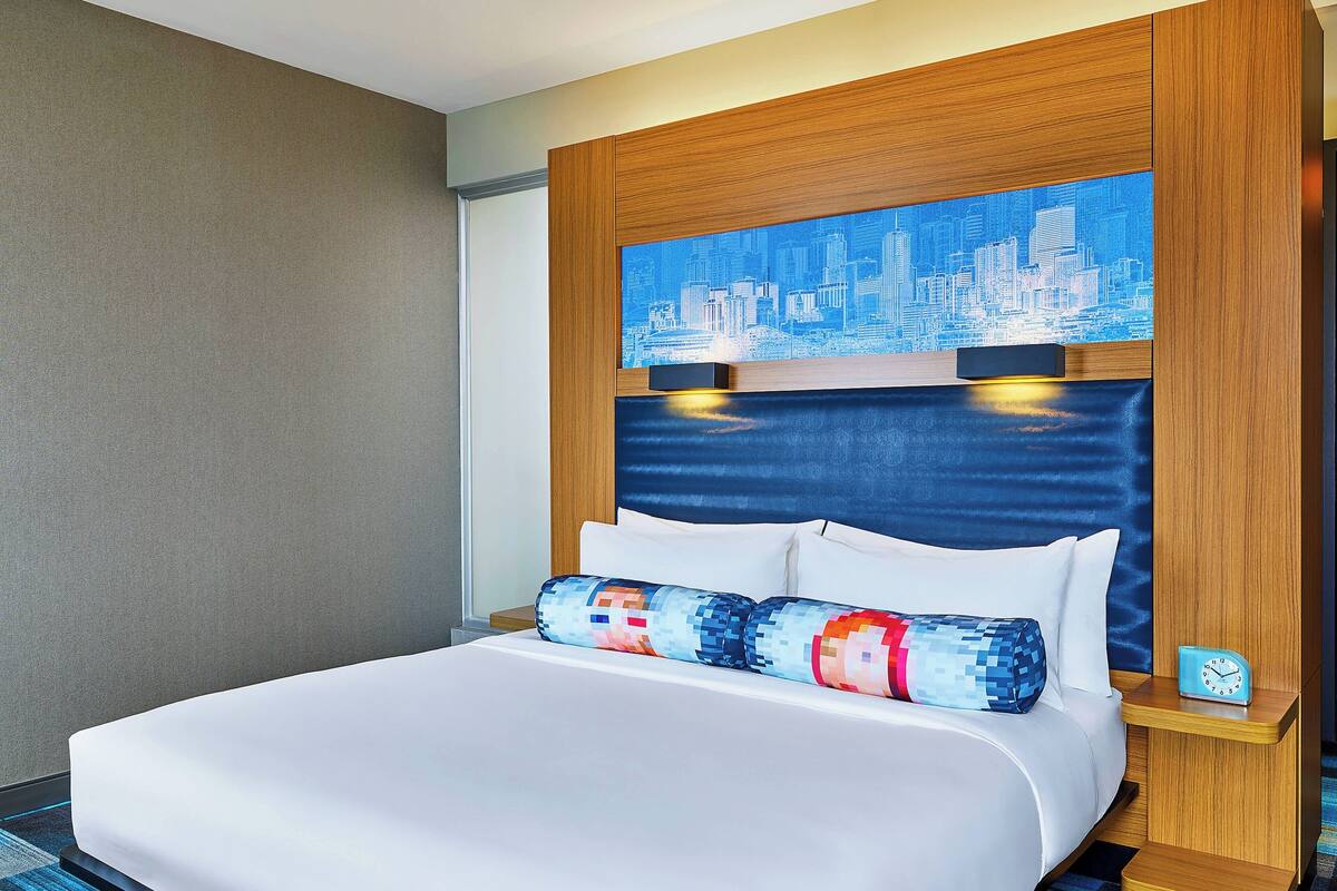 A hotel room with a large bed, a cityscape artwork above the headboard, and a small clock on the nightstand.