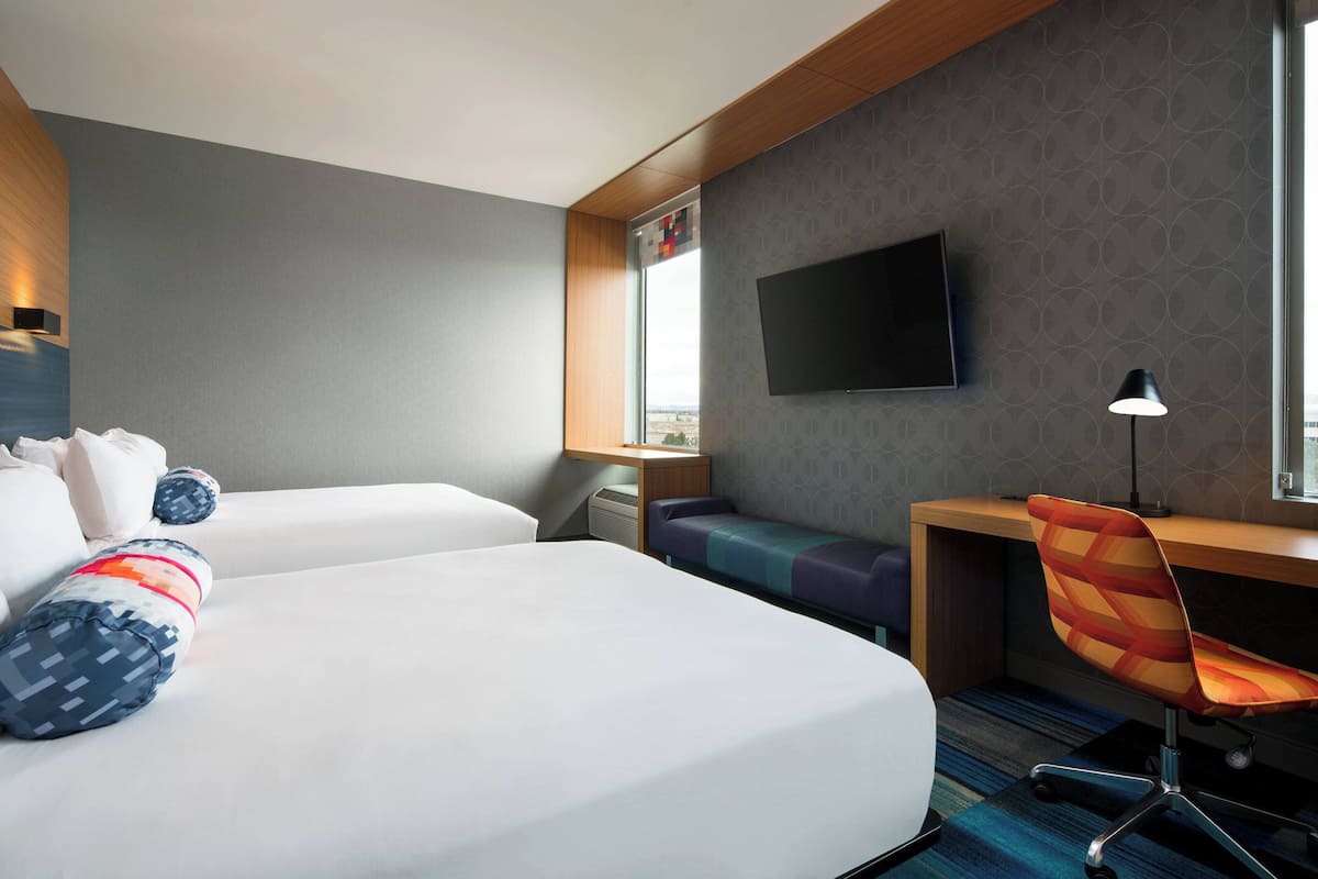 A hotel room with two beds, a desk, a chair, a flat-screen TV, and a window.