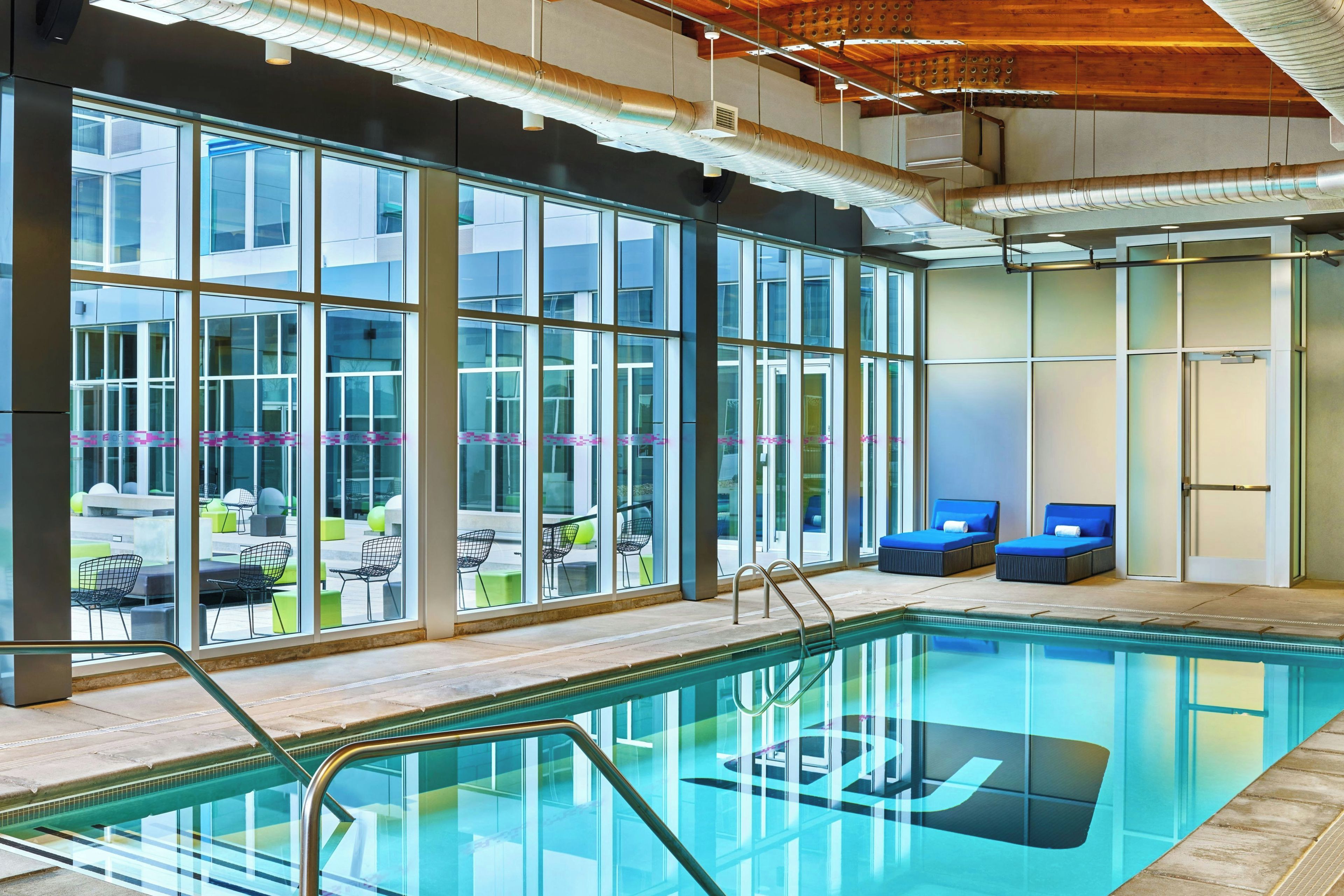 Una piscina interior moderna con paredes de vidrio transparente, sillas reclinables azules y vistas a un área de estar exterior.