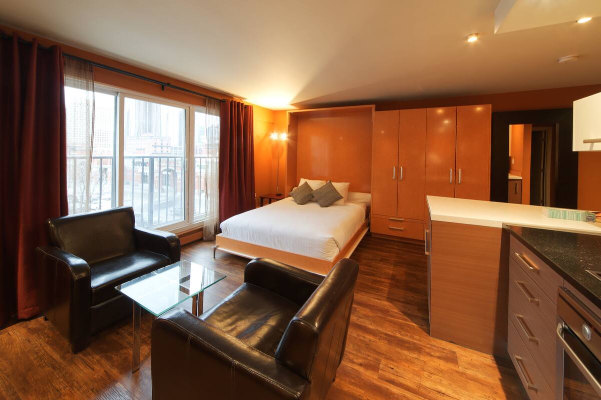 A modern hotel room with a bed, a glass coffee table, a leather sofa, wooden cabinets, and a view of the city.