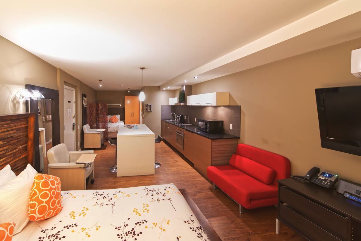 A modern hotel room with a bed, a red sofa, a kitchen area, and a TV mounted on the wall.