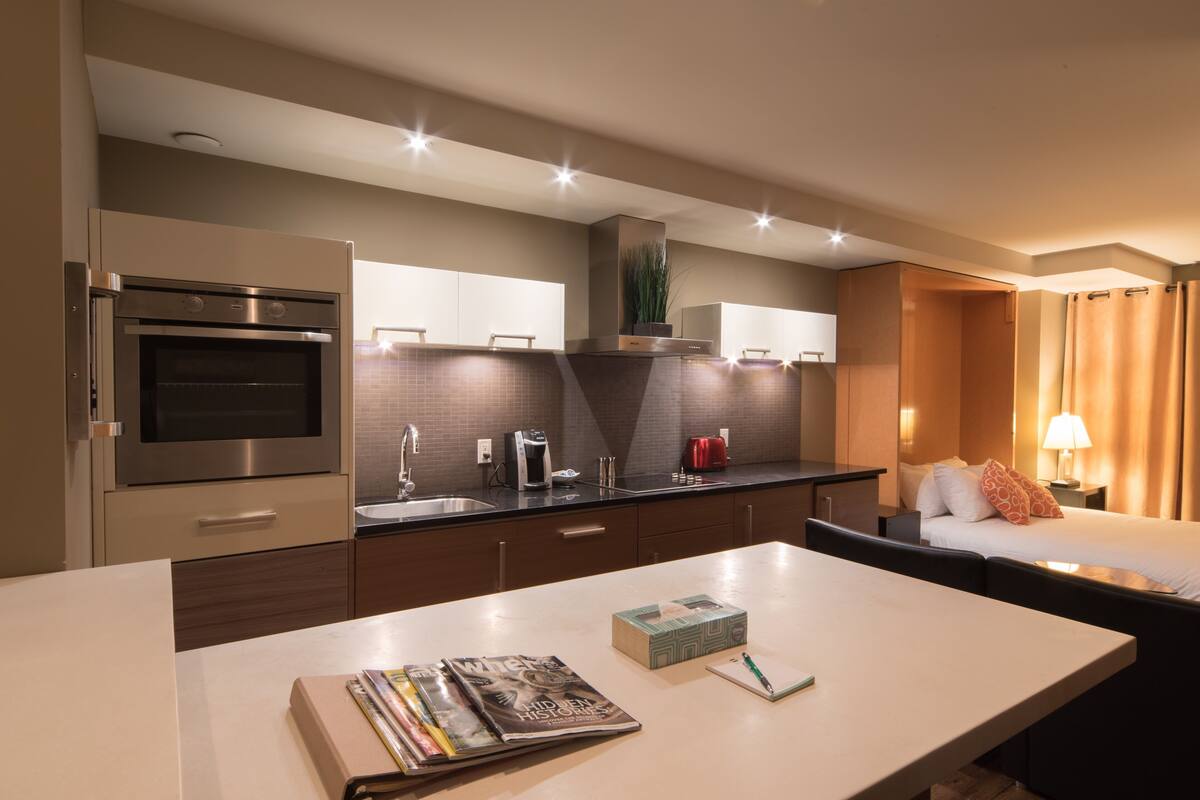 A modern hotel kitchen with a built-in oven, a coffee machine, and a sink. There is a dining table with magazines and a notebook.