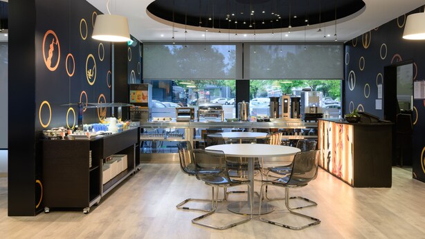 A modern cafe with a central round table, clear chairs, a counter with a menu, and a large window offering a view of the street. - Star Inn Porto (Porto)