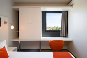 A hotel room with a bed, a desk with a chair, a wardrobe, and a window with a view of buildings.