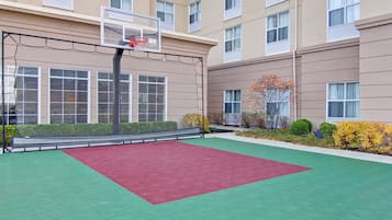 An indoor basketball court with a red and green floor, a basketball hoop, and a building with multiple windows in the background.