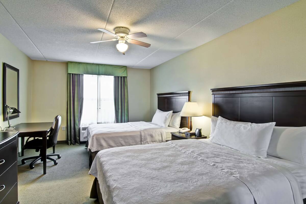 Une chambre d’hôtel avec deux lits, un bureau, une chaise, un ventilateur de plafond et une fenêtre avec des rideaux.