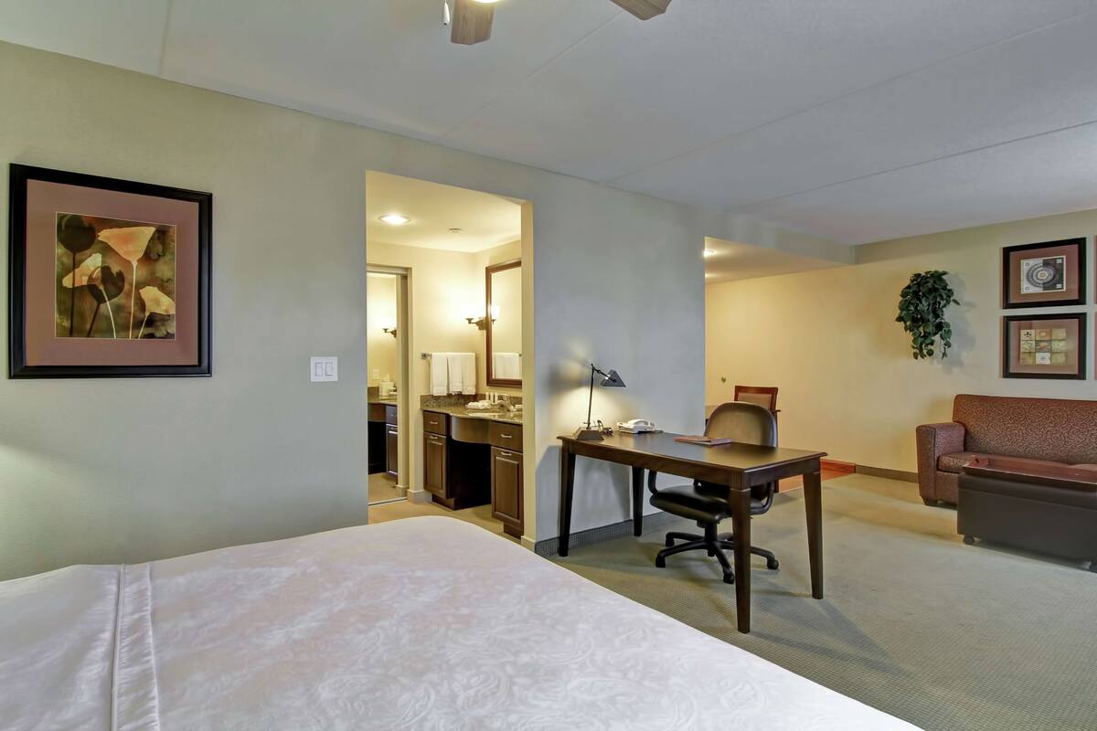 Une chambre d’hôtel avec un lit, un bureau, une chaise et un petit coin salon. On aperçoit une kitchenette au fond de la pièce.